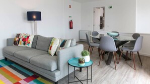 Un salon moderne avec un canapé gris, des coussins colorés, une petite table ronde ornée d’une plante, et un coin repas avec des chaises grises et une table en verre.