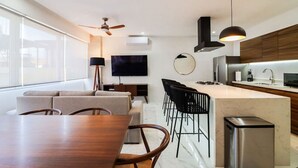A modern kitchen with a central island, a dining area with a wooden table and chairs, a sofa, and a TV mounted on the wall.