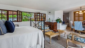 Een ruime kamer met een bed, een houten stoel, een klein tafeltje en een bureau met een stoel. Er is een raam met uitzicht op groen.