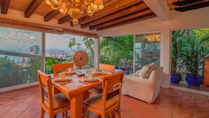 A dining area with a wooden table, chairs, and a chandelier. There are large windows with a view of greenery and a building in the background. - Studio La Vista – Stunning Views in the Heart of Puerto Vallarta (Puerto Vallarta)