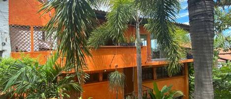 A multi-story building with orange walls and balconies, surrounded by lush greenery and palm trees.