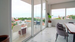 A spacious balcony with large windows offering a city view, a dining area with chairs and a table, and a potted plant.