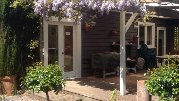 A wooden house with a covered patio, a small garden with potted plants, and a pergola with purple flowers.