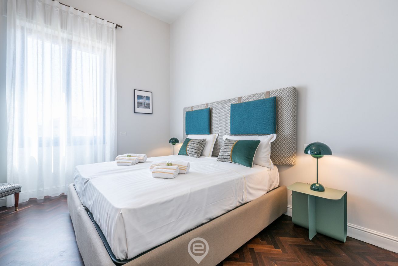 A neatly made bed with white linens and multiple pillows, a bedside table with a lamp, and a window with sheer curtains.