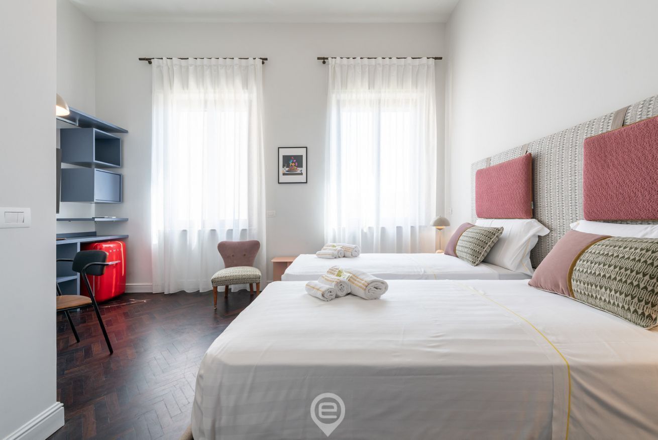 A hotel room with two beds, a desk with a chair, a red suitcase, and a picture on the wall.