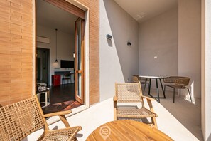 Un patio exterior iluminado por el sol con mobiliario de mimbre y una mesa de madera, que da acceso a un espacio interior con cocina y comedor.