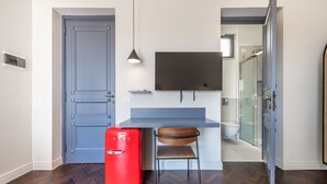 Une pièce de taille modeste avec une porte bleue, un petit réfrigérateur rouge, une chaise en bois, un téléviseur à écran plat fixé au mur et un espace douche visible à travers une porte en verre.