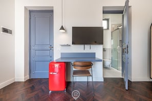 A compact room with a blue door, a small red refrigerator, a wooden chair, a mounted flat-screen TV, and a shower area visible through a glass door.