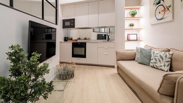 A modern kitchen with white cabinets, a microwave, and a stove. There is a beige sofa with patterned cushions and a small plant on the floor. - Charming Almudena I - (Madrid)
