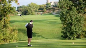 Un campo de golf con un jugador a punto de golpear, árboles y cielo despejado.