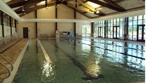 Una piscina interior con carriles delimitados, vigas de madera en el techo y grandes ventanales en un lateral.