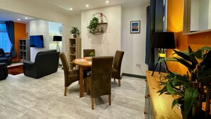 Ein modernes Wohnzimmer mit Essbereich, ausgestattet mit einem Holztisch und Stühlen, einem Bücherregal, einem Fernseher und einer Pflanze.