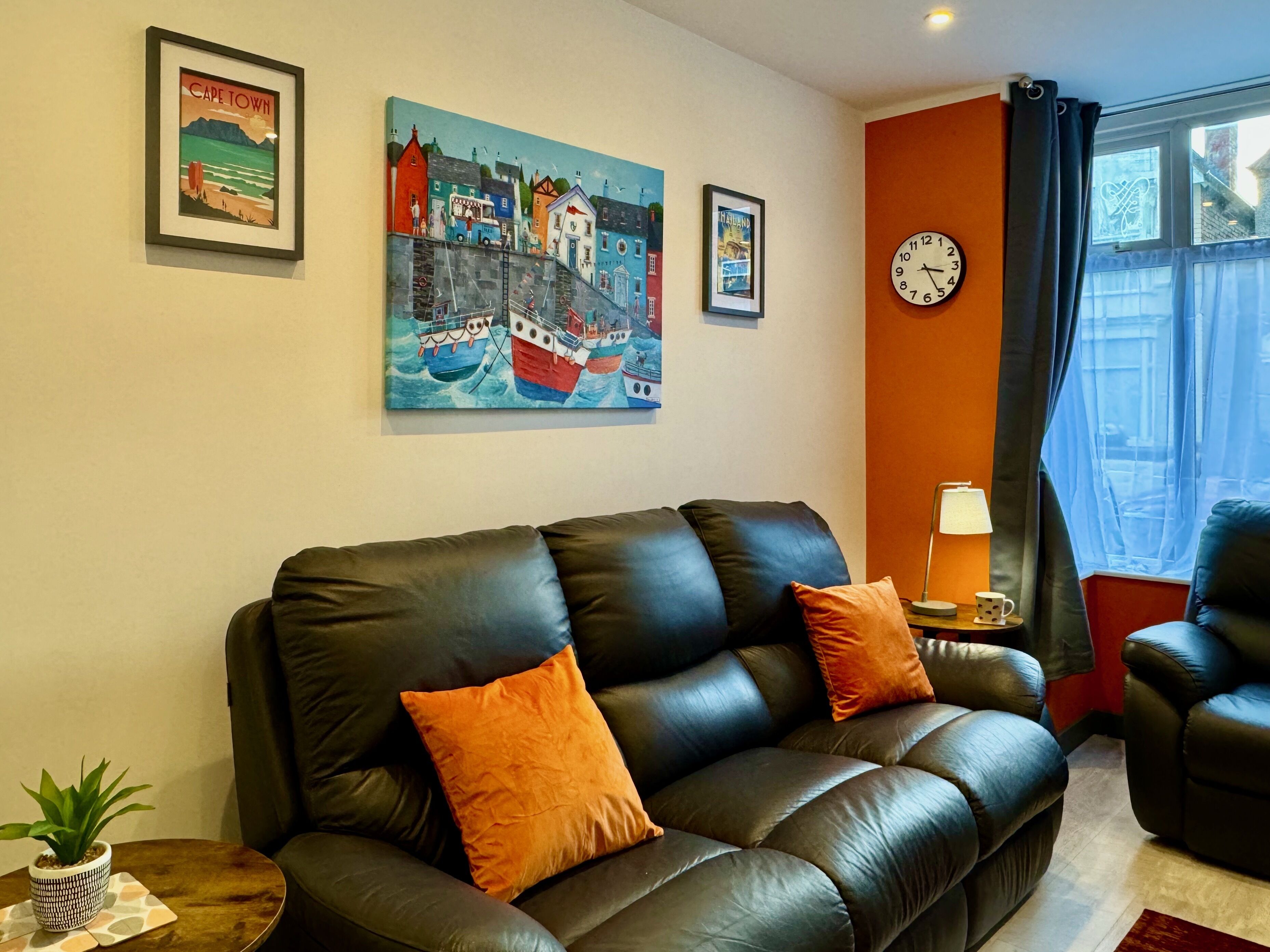 A living room with a black leather sofa, two orange cushions, a small table with a plant, and a lamp. There are framed pictures on the wall, including one labeled "CAPE TOWN" and another with a boat. A clock is visible on the wall, and a window with curtains is on the right.
