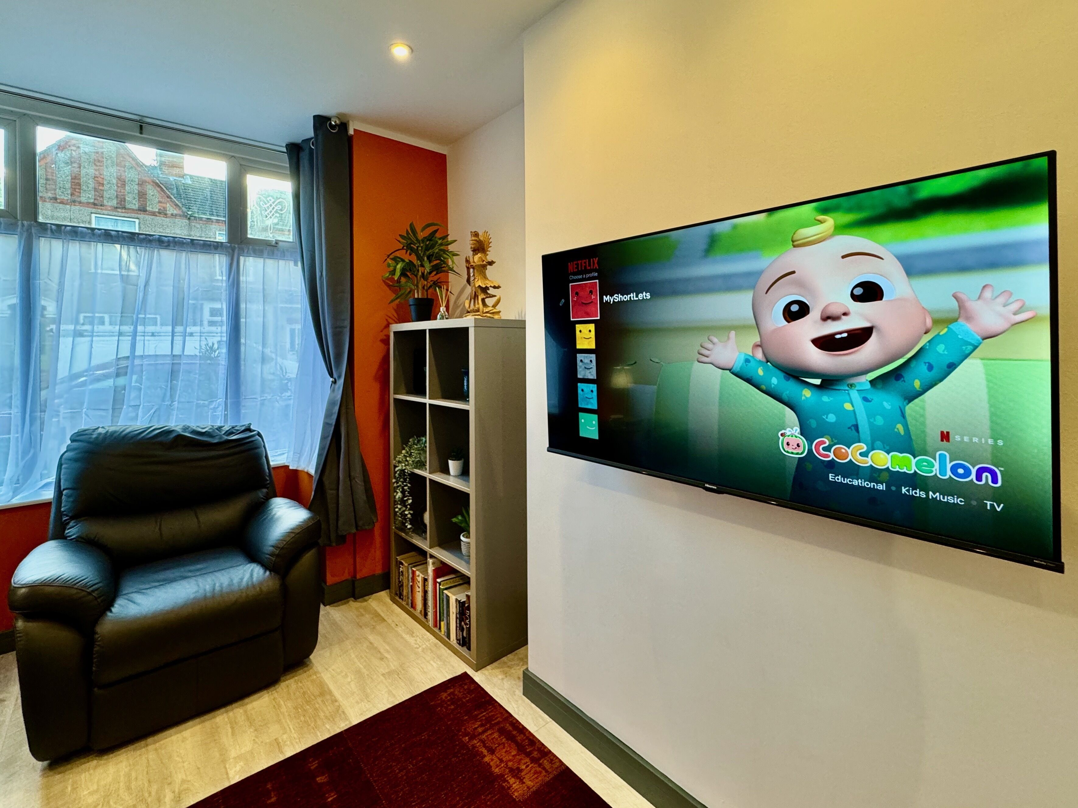 A living room with a black leather sofa, a bookshelf, a TV displaying 'Cocomelon', and a window with curtains.