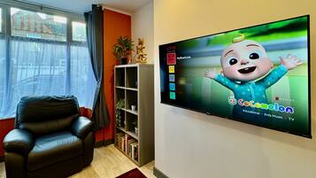 A living room with a black leather sofa, a bookshelf, a TV displaying 'Cocomelon', and a window with curtains.