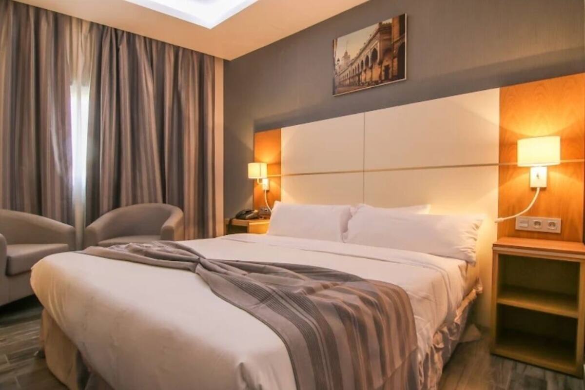 A hotel room with a large bed, two bedside lamps, a nightstand, a chair, and a painting on the wall.