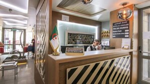 A hotel reception area with a modern design, featuring a reception desk, a flag, and a sign indicating room types. - El Ghanami Hotel (Algiers)