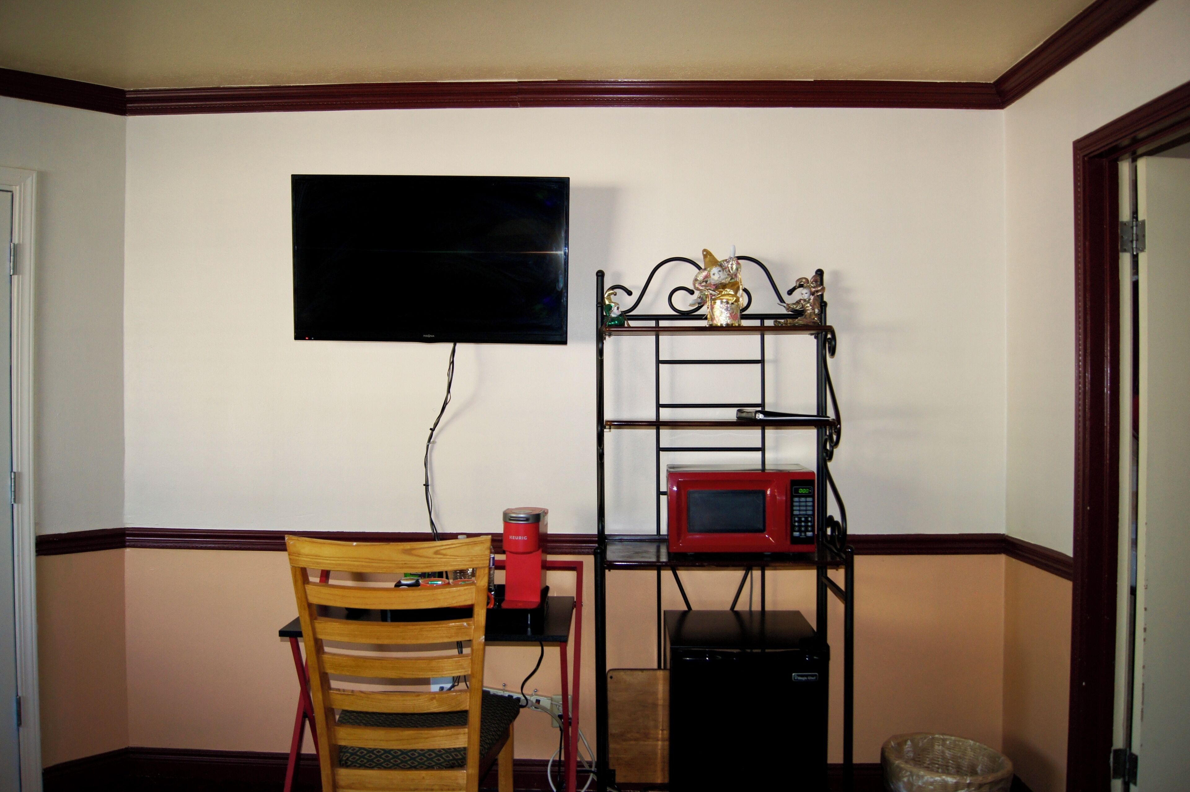 A small room with a television mounted on the wall, a red coffee maker on a table, a microwave on a shelf, and a wicker basket on the floor.