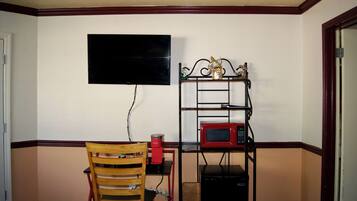A small room with a television mounted on the wall, a red coffee maker on a table, a microwave on a shelf, and a wicker basket on the floor.