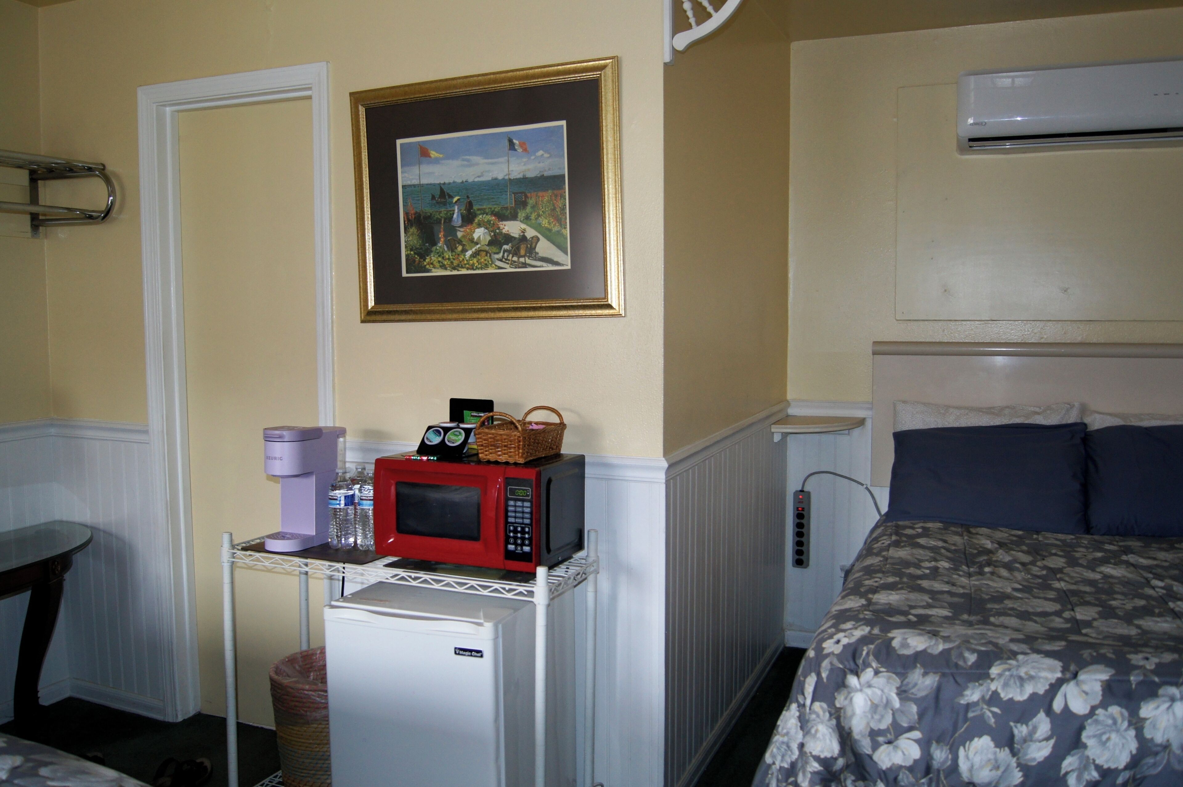 Una habitación compacta con cama, una kitchenette pequeña con microondas y un cuadro enmarcado en la pared.