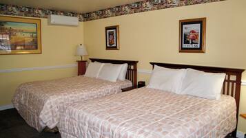 A hotel room with two beds, a wall-mounted air conditioner, a lamp, and framed pictures on the wall.