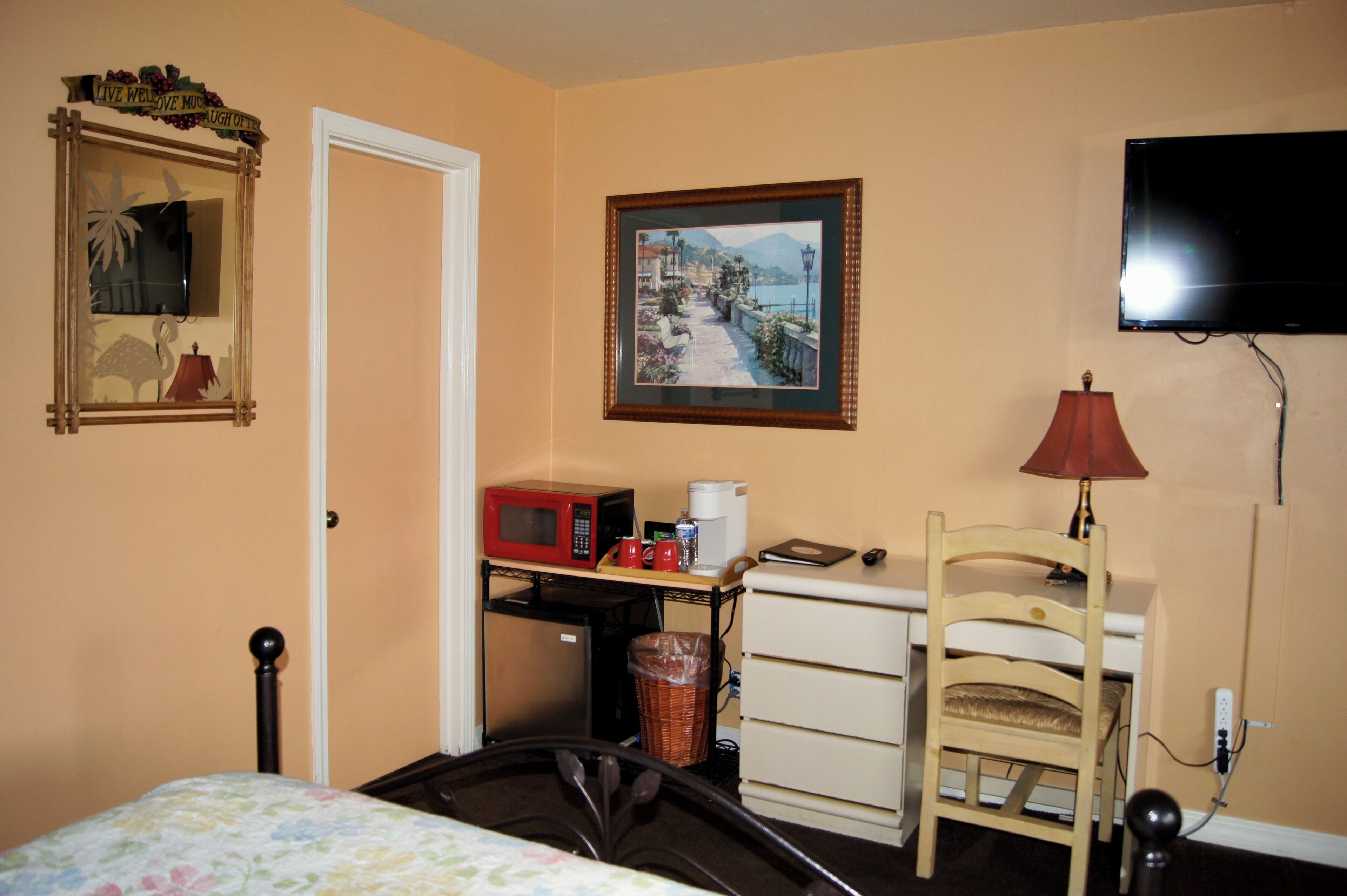 Quarto de hotel com cama, escrivaninha com micro-ondas, cadeira, lixeira, cômoda, televisão e um quadro na parede.