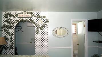 A bedroom with a bed, a wall-mounted TV, a door leading to a bathroom, and a sign reading "WELCOME TO OUR GARDEN".