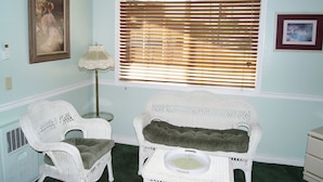 Una habitación con dos sillas blancas de mimbre, una lámpara de pie, una ventana con persianas y un cuadro en la pared.