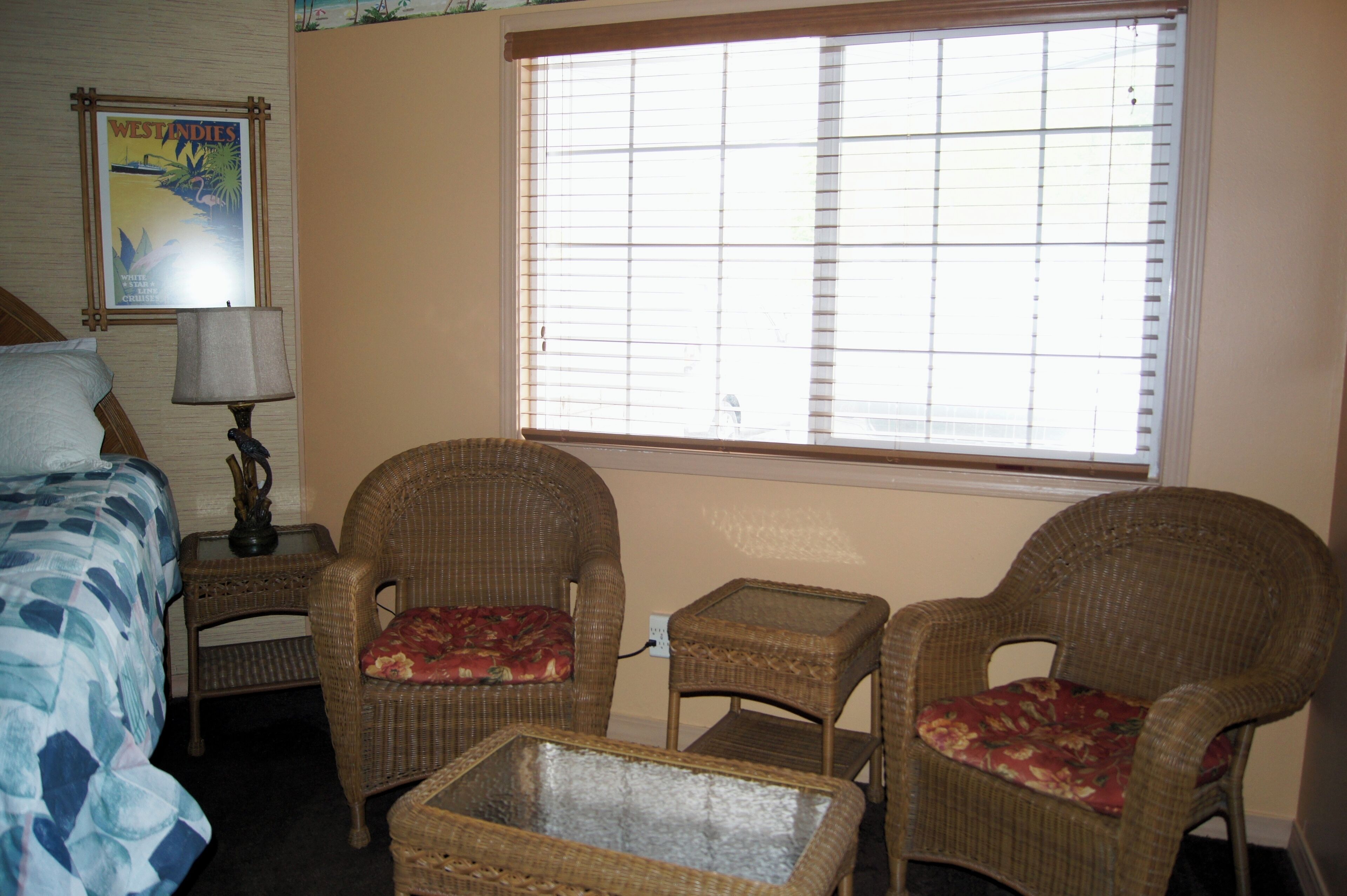 A hotel room with a bed, two wicker chairs with floral cushions, a small table, a lamp, and a window with blinds.