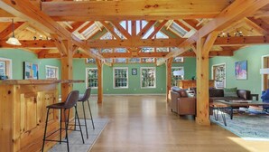 Une pièce spacieuse avec un haut plafond en bois, un coin bar avec des tabourets, un espace salon avec un canapé et des fauteuils, et de grandes fenêtres laissant entrer la lumière naturelle.