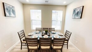 A dining room with a round table set for six, flanked by two windows with blinds, and framed abstract art on the walls.