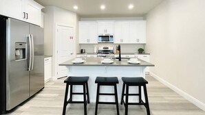 A modern kitchen with white cabinets, a stainless steel refrigerator, a microwave, and a central island with bar stools.