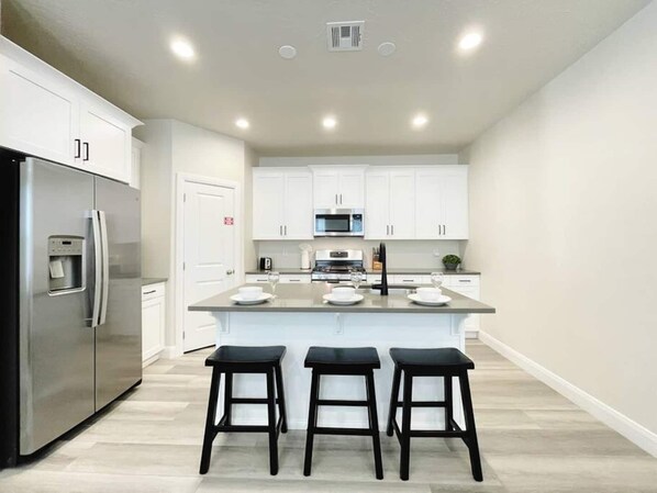 A modern kitchen with white cabinets, a stainless steel refrigerator, a microwave, and a central island with bar stools.