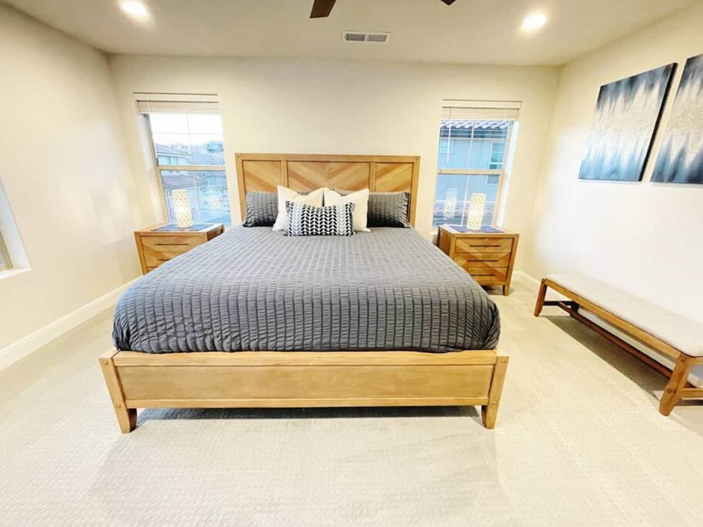 A bedroom with a large bed, two bedside tables, a bench, and two windows with blinds.