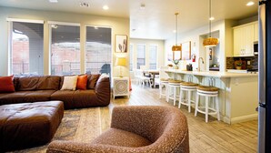 A modern living room with a brown leather sofa, a patterned armchair, and a dining area with white chairs and a kitchen island.