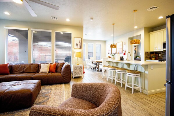 A modern living room with a brown leather sofa, a patterned armchair, and a dining area with white chairs and a kitchen island.