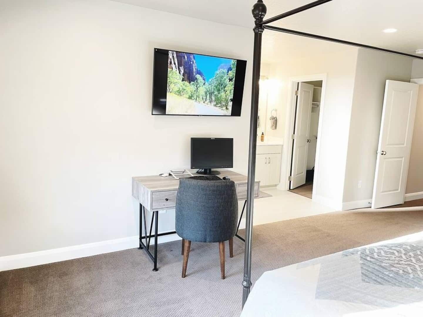 Une chambre à coucher comprenant un lit, un bureau avec un ordinateur, un téléviseur fixé au mur et une porte donnant accès à une salle de bain.
