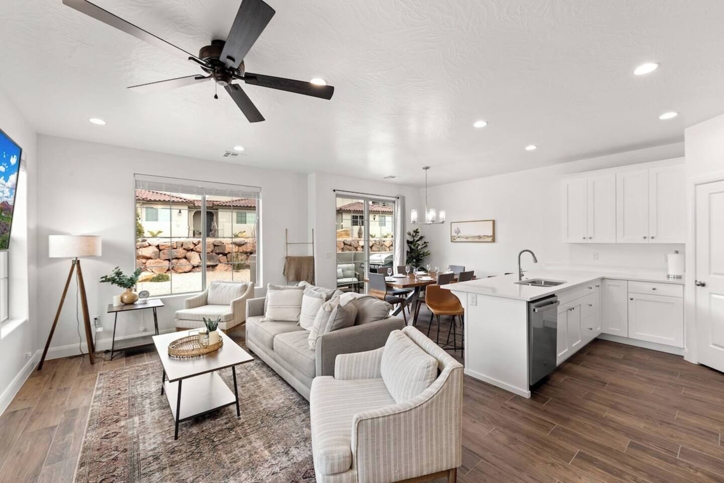 A spacious living area with a sofa, coffee table, and dining table, a kitchen with white cabinets, and a ceiling fan.