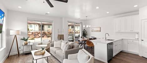 A spacious living area with a sofa, coffee table, and dining table, a kitchen with white cabinets, and a ceiling fan.