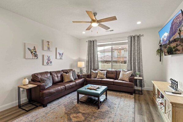 A living room with a brown leather sectional sofa, a coffee table, a ceiling fan, and a large window with curtains. - Southern Utah charm. Close to St George and National Parks (Washington)