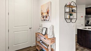 A modern hallway with a white door, a wooden console table with decorative items, a geometric wall shelf, and a kitchen area in the background. - Southern Utah charm. Close to St George and National Parks (Washington)