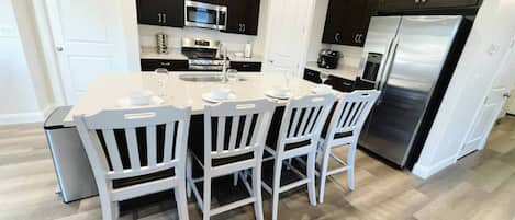 A modern kitchen with dark cabinets, a stainless steel refrigerator, and a central island with white chairs.
