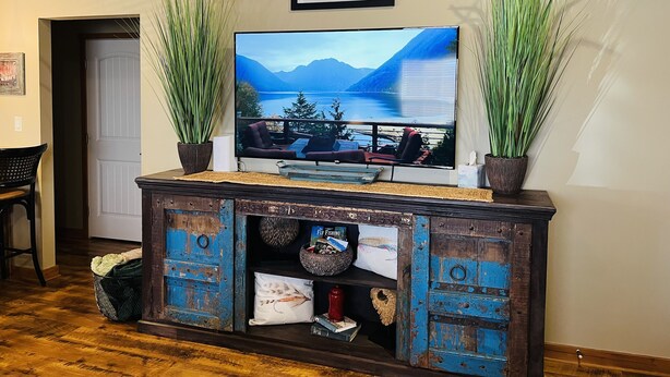 Un salon doté d’un grand téléviseur à écran plat fixé sur un meuble en bois rustique, de part et d’autre duquel se trouvent de grandes plantes vertes.