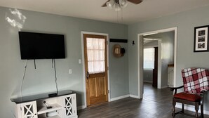 A living room with a TV mounted on the wall, a wooden cabinet, a door with a window, a chair with a blanket, and a hallway leading to another room. - Cozy cottage in delightful Ahwahnee with AC, WiFi (Ahwahnee)