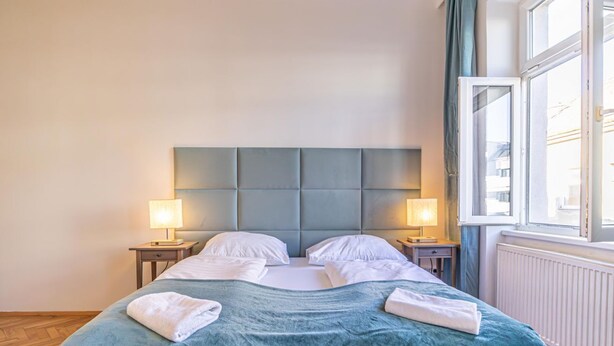 A neatly made bed with a blue bedspread and white pillows, two bedside lamps, and a window with curtains.