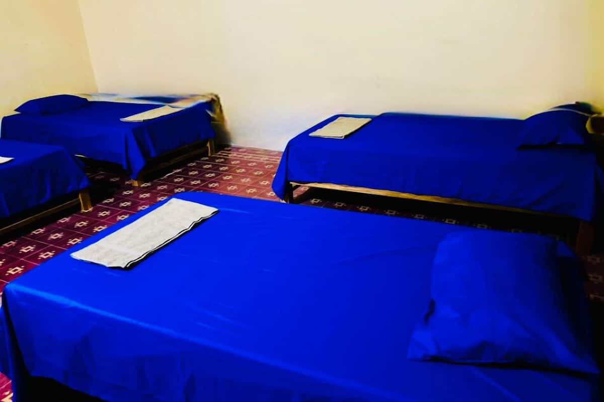 Ein Zimmer mit vier Einzelbetten, jeweils mit blauen Laken und Kissen bezogen. Der Raum verfügt über einen gemusterten Teppich und schlichte Wände.