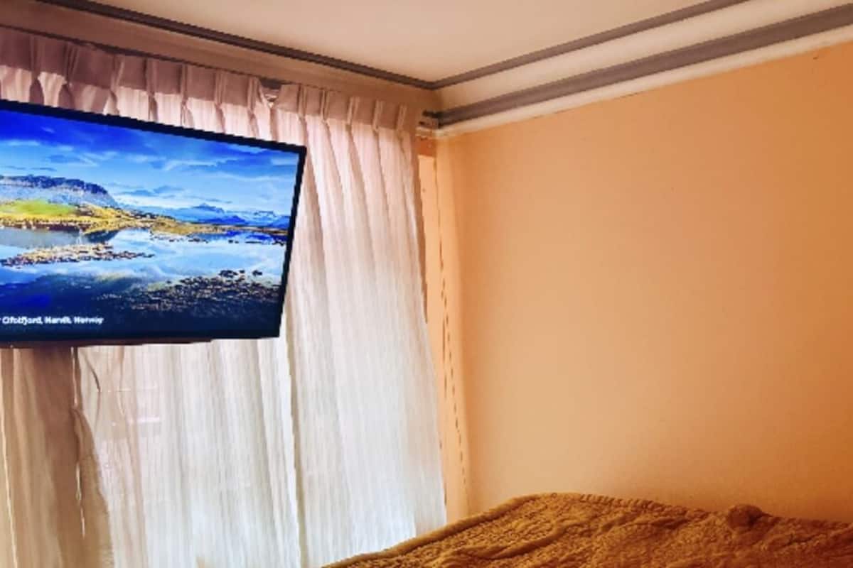 Ein Zimmer mit einem an der Wand befestigten Fernseher, einem Bett mit brauner Decke und einem Fenster mit transparenten Vorhängen.