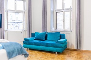 Quarto com sofá azul, piso de madeira e grandes janelas com cortinas.