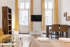 Hotelkamer met een bed, een glazen bijzettafeltje, een aan de muur bevestigde televisie, een houten boekenkast, een eettafel met stoelen en grote ramen met gordijnen.