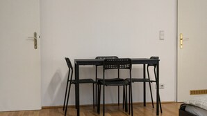 Une pièce avec un plancher en bois, une table noire et quatre chaises, un lit recouvert d’une couverture grise et des murs blancs.